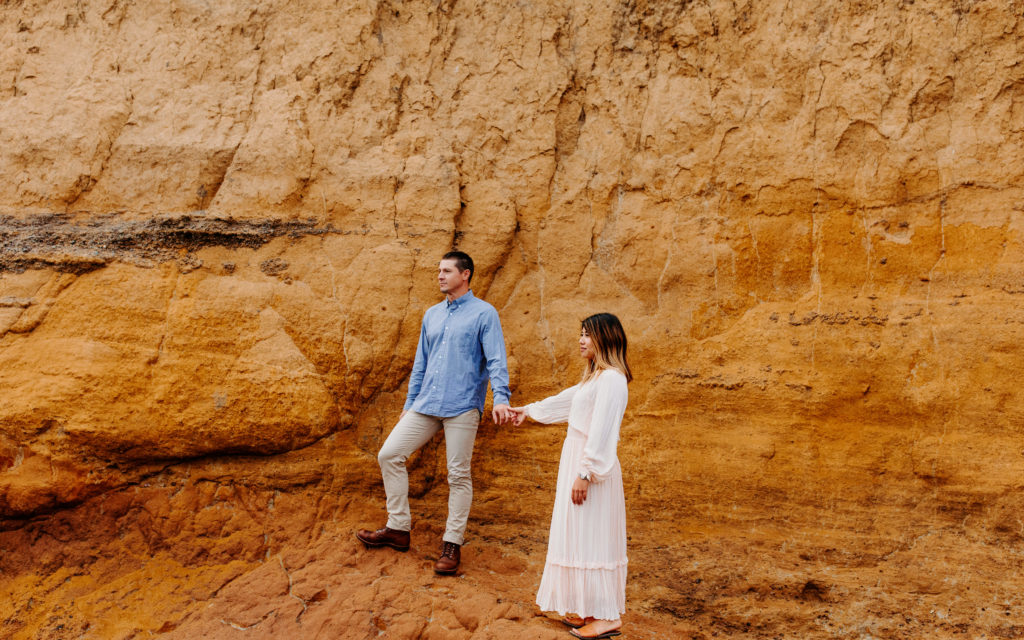 couple holding hands on orange rock on beach, Marin Headlands Engagement Photos, Bay Area Engagement Photos, Bay Area Engagement Photo Locations, Engagement Photo Outfits, Bay Area Beach Engagement Photos, Dog Friendly Bay Area Beaches, Best Locations for Bay Area Engagement Photos, San Francisco Engagement Photos, What to Wear for Engagement Photos, Engagement Photos with dogs, Rachel Christopherson Photography LLC