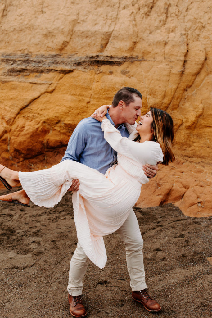 man cradling woman while laughing, Marin Headlands Engagement Photos, Bay Area Engagement Photos, Bay Area Engagement Photo Locations, Engagement Photo Outfits, Bay Area Beach Engagement Photos, Dog Friendly Bay Area Beaches, Best Locations for Bay Area Engagement Photos, San Francisco Engagement Photos, What to Wear for Engagement Photos, Engagement Photos with dogs, Rachel Christopherson Photography LLC