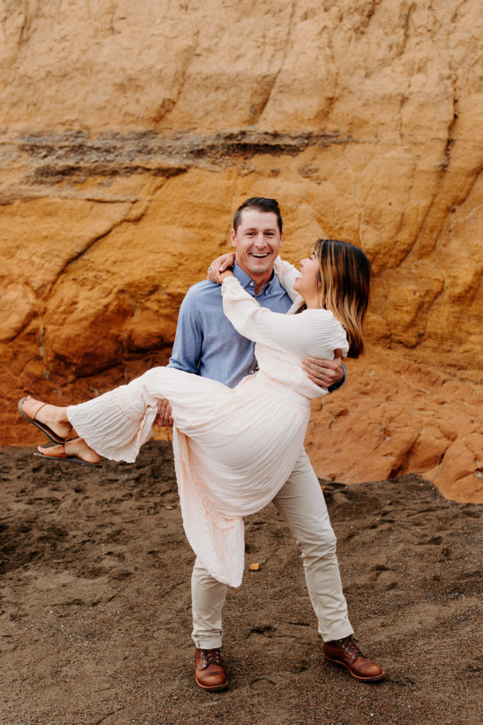 man cradling woman on orange rock, Marin Headlands Engagement Photos, Bay Area Engagement Photos, Bay Area Engagement Photo Locations, Engagement Photo Outfits, Bay Area Beach Engagement Photos, Dog Friendly Bay Area Beaches, Best Locations for Bay Area Engagement Photos, San Francisco Engagement Photos, What to Wear for Engagement Photos, Engagement Photos with dogs, Rachel Christopherson Photography LLC