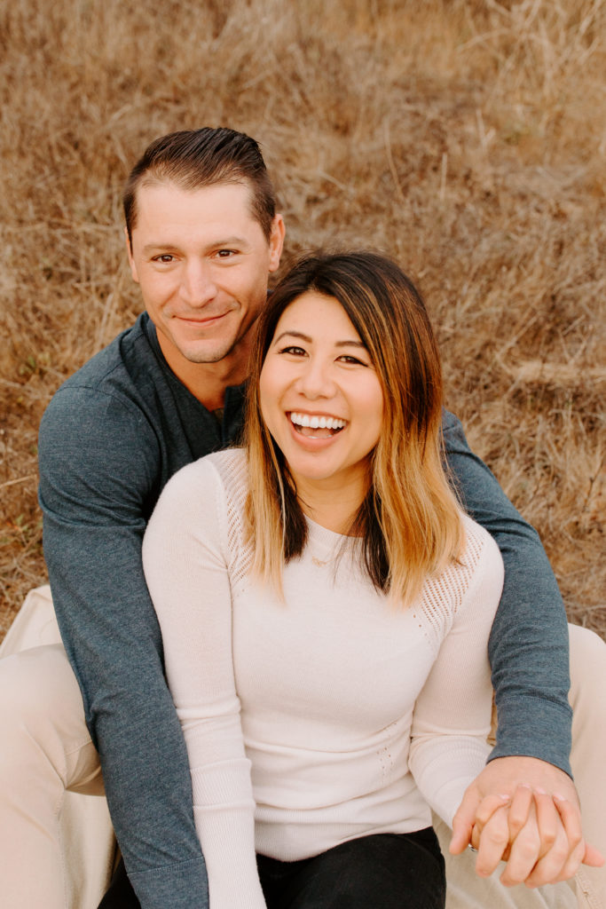 couple sitting and smiling at camera in grass, Marin Headlands Engagement Photos, Bay Area Engagement Photos, Bay Area Engagement Photo Locations, Engagement Photo Outfits, Coastal Bay Area Engagement Photos, Dog Friendly Bay Area Beaches, Best Locations for Bay Area Engagement Photos, San Francisco Engagement Photos, What to Wear for Engagement Photos, Engagement Photos with dogs, Rachel Christopherson Photography LLC