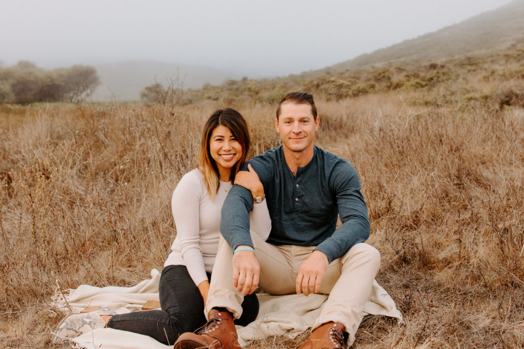 couple smiling at camera sitting in field, Marin Headlands Engagement Photos, Bay Area Engagement Photos, Bay Area Engagement Photo Locations, Engagement Photo Outfits, Coastal Bay Area Engagement Photos, Dog Friendly Bay Area Beaches, Best Locations for Bay Area Engagement Photos, San Francisco Engagement Photos, What to Wear for Engagement Photos, Engagement Photos with dogs, Rachel Christopherson Photography LLC