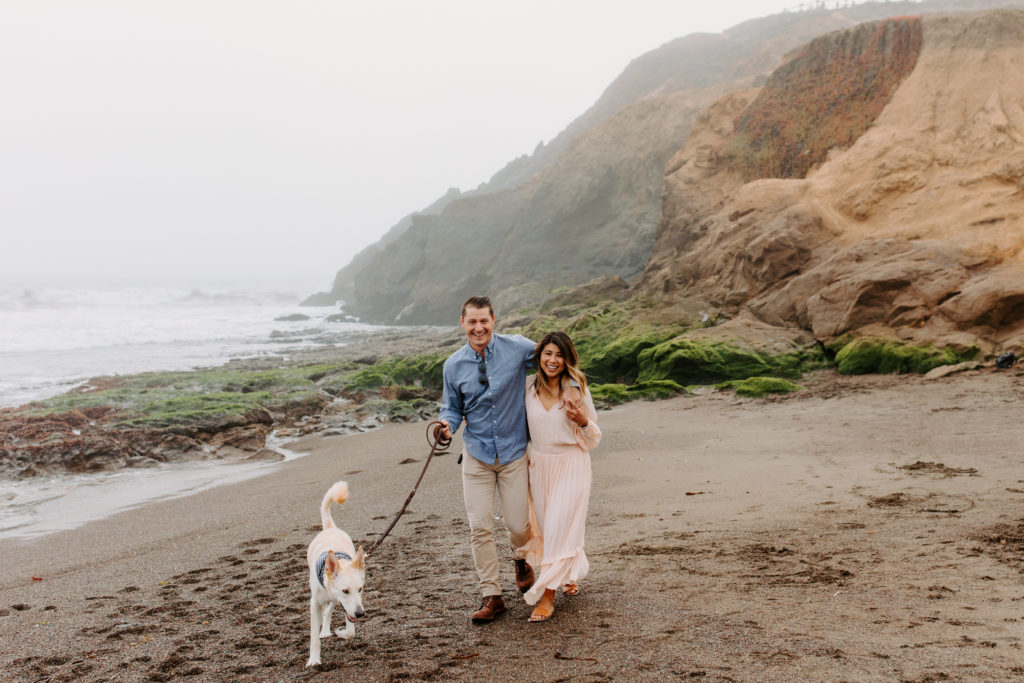 couple smiling at camera walking dog, Marin Headlands Engagement Photos, Bay Area Engagement Photos, Bay Area Engagement Photo Locations, Engagement Photo Outfits, Bay Area Beach Engagement Photos, Dog Friendly Bay Area Beaches, Best Locations for Bay Area Engagement Photos, San Francisco Engagement Photos, What to Wear for Engagement Photos, Engagement Photos with dogs, Rachel Christopherson Photography LLC