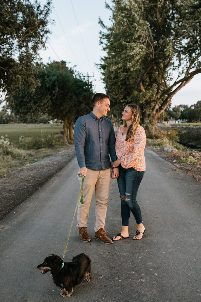 Sacramento River Engagement Session Northern California Weiner dog engagement shoot outfit inspo engagement photos with dog couples shoot posing golden hour sacramento river rachel christopherson photography tree lined street walk holding hands