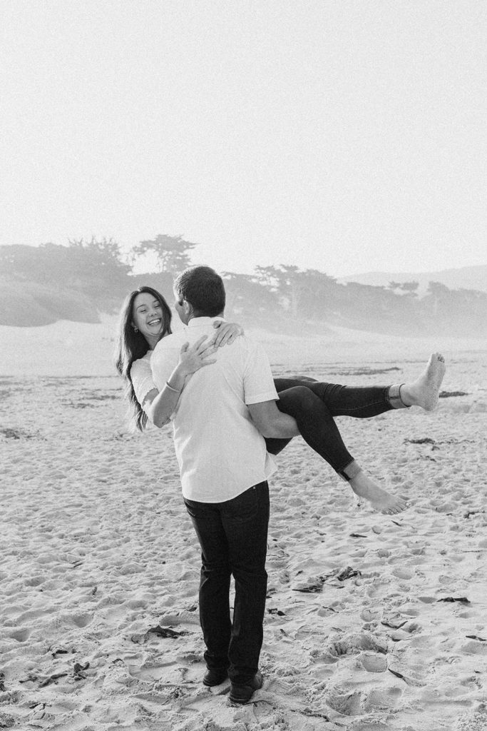 black and white photo, man lifting woman playfully on beach, Carmel CA Engagement Photos, Monterey Engagement Photos, Monterey Engagement Photo Locations, Engagement Photo Outfits, California Beach Engagement Photos, Best Locations for Monterey Engagement Photos, What to Wear for Engagement Photos, Rachel Christopherson Photography LLC