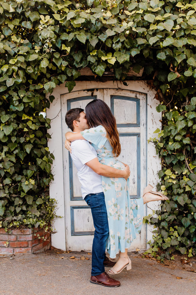 man lifts woman in front of vine covered door, Carmel CA Engagement Photos, Monterey Engagement Photos, Monterey Engagement Photo Locations, Engagement Photo Outfits, Carmel by the sea Engagement Photos, Best Locations for Monterey Engagement Photos, What to Wear for Engagement Photos, Rachel Christopherson Photography LLC