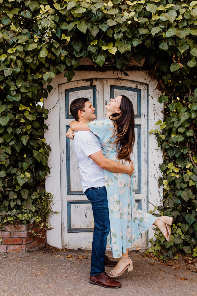 man lifts woman in front of vine covered door, Carmel CA Engagement Photos, Monterey Engagement Photos, Monterey Engagement Photo Locations, Engagement Photo Outfits, Carmel by the sea Engagement Photos, Best Locations for Monterey Engagement Photos, What to Wear for Engagement Photos, Rachel Christopherson Photography LLC