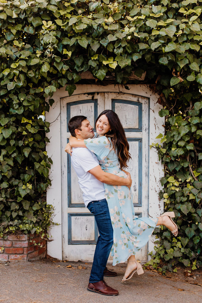 man lifts woman in front of vine covered door, Carmel CA Engagement Photos, Monterey Engagement Photos, Monterey Engagement Photo Locations, Engagement Photo Outfits, Carmel by the sea Engagement Photos, Best Locations for Monterey Engagement Photos, What to Wear for Engagement Photos, Rachel Christopherson Photography LLC