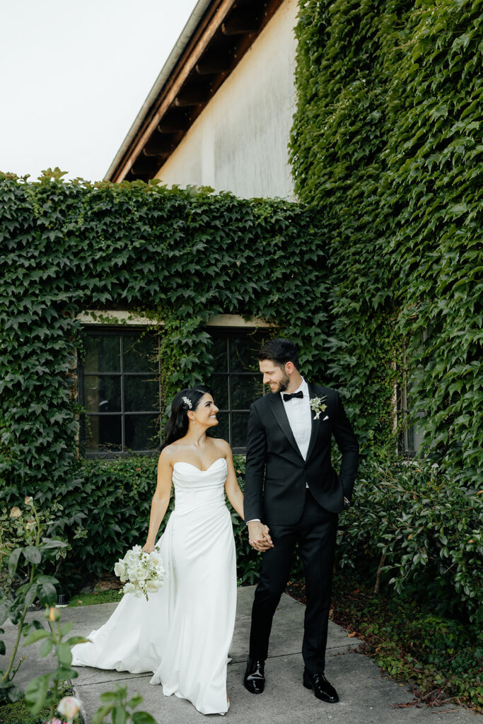 Clos LaChance Wedding - Bride and groom holding hands walking with next to vine covered bricks